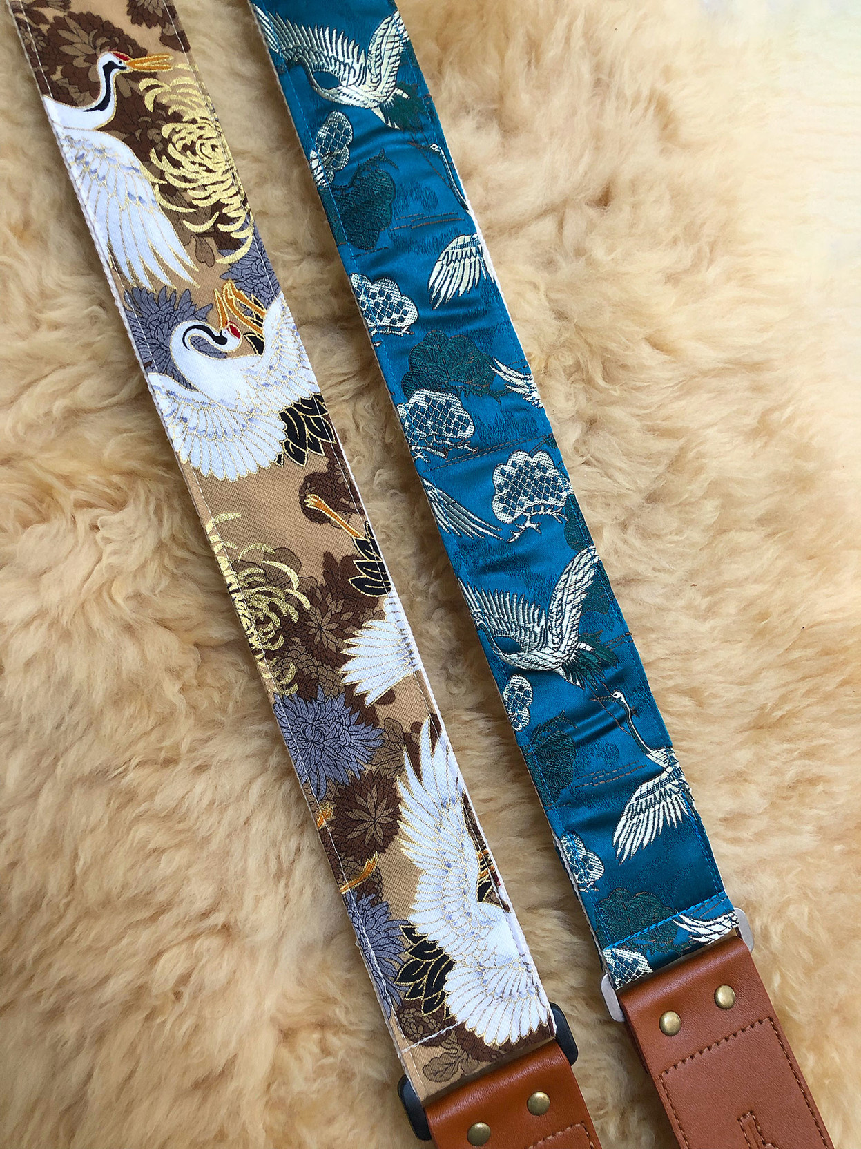 Two guitar straps, each featuring flying cranes. On the left is matte cloth with tans and earth tones, while on the right is blue satin with gold and green.