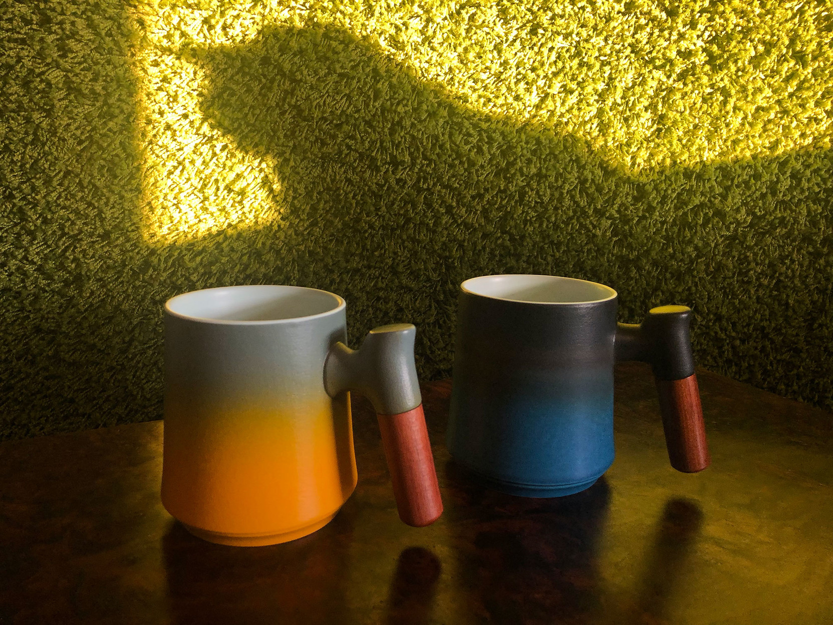 Two teacups with wooden handles – one orange and grey, the other black fading to dark aqua blue – sit in the foreground,

while the shadow of Milla the kitty looms over them, cast on the grass green shag carpet hanging on the wall in the background.