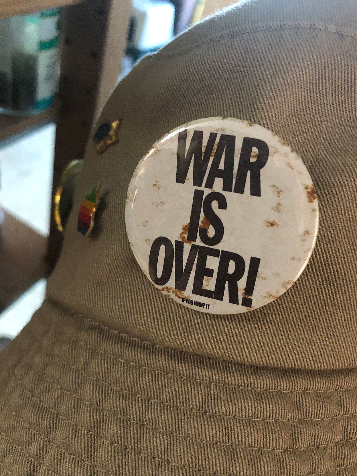 Closeup of a khaki hat with a slightly rusty “War is Over” button attached.