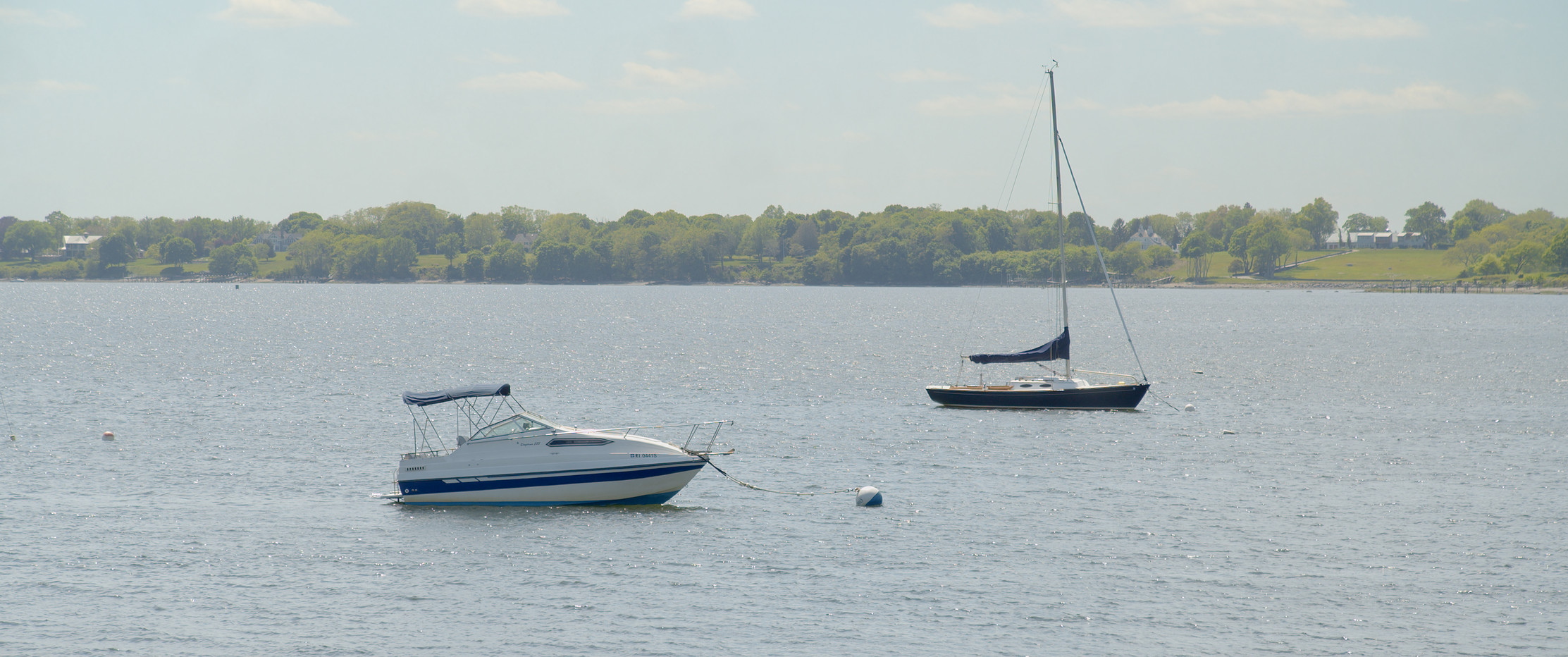 A sunny, hazy spring day looking out into the bay.

Two sailboats at anchor in Bristol Harbor, with the green arc of trees on Poppasquash Point in the background.
