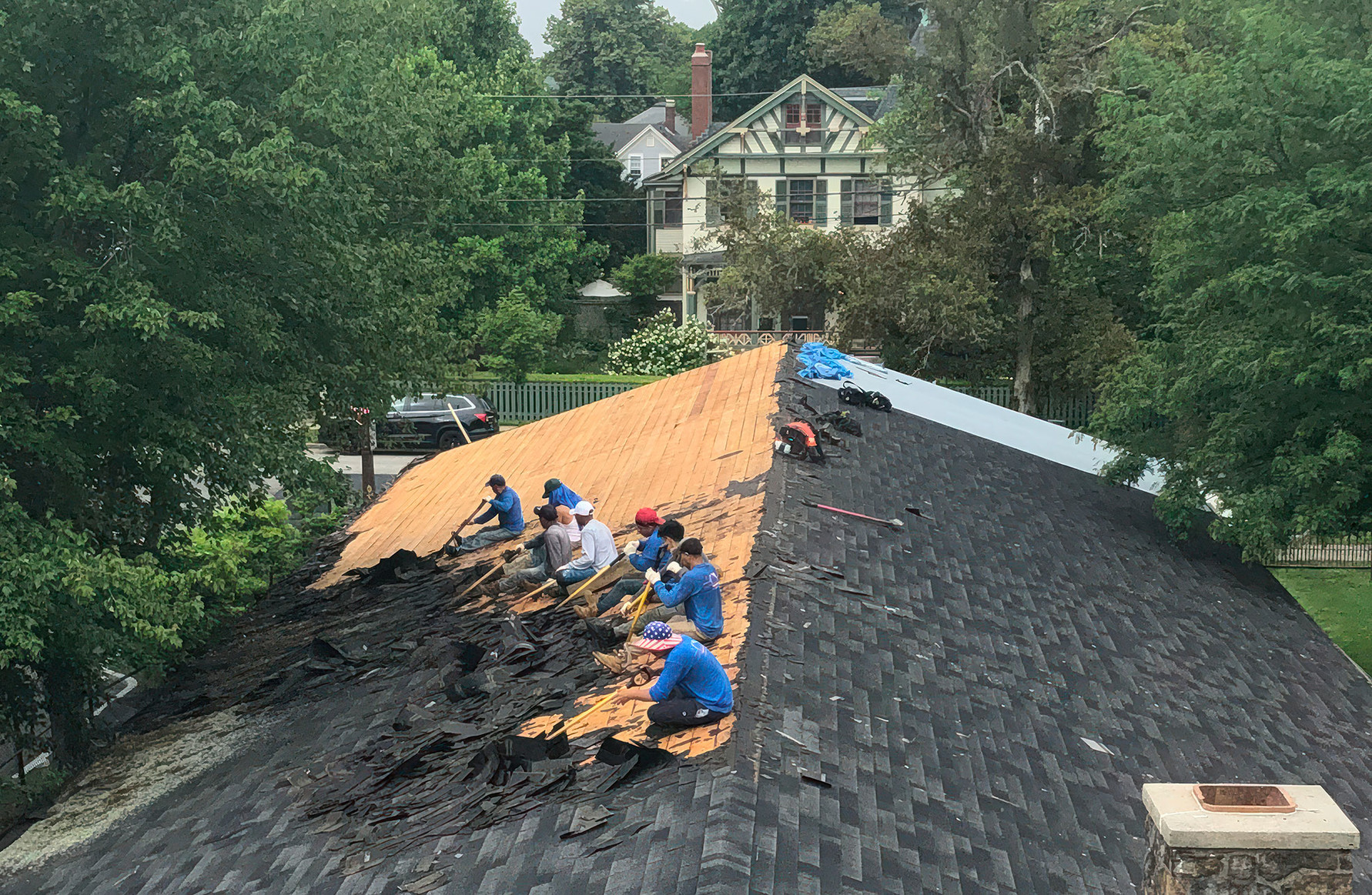A crew of workers removing shingles from the roof of a church building next door.