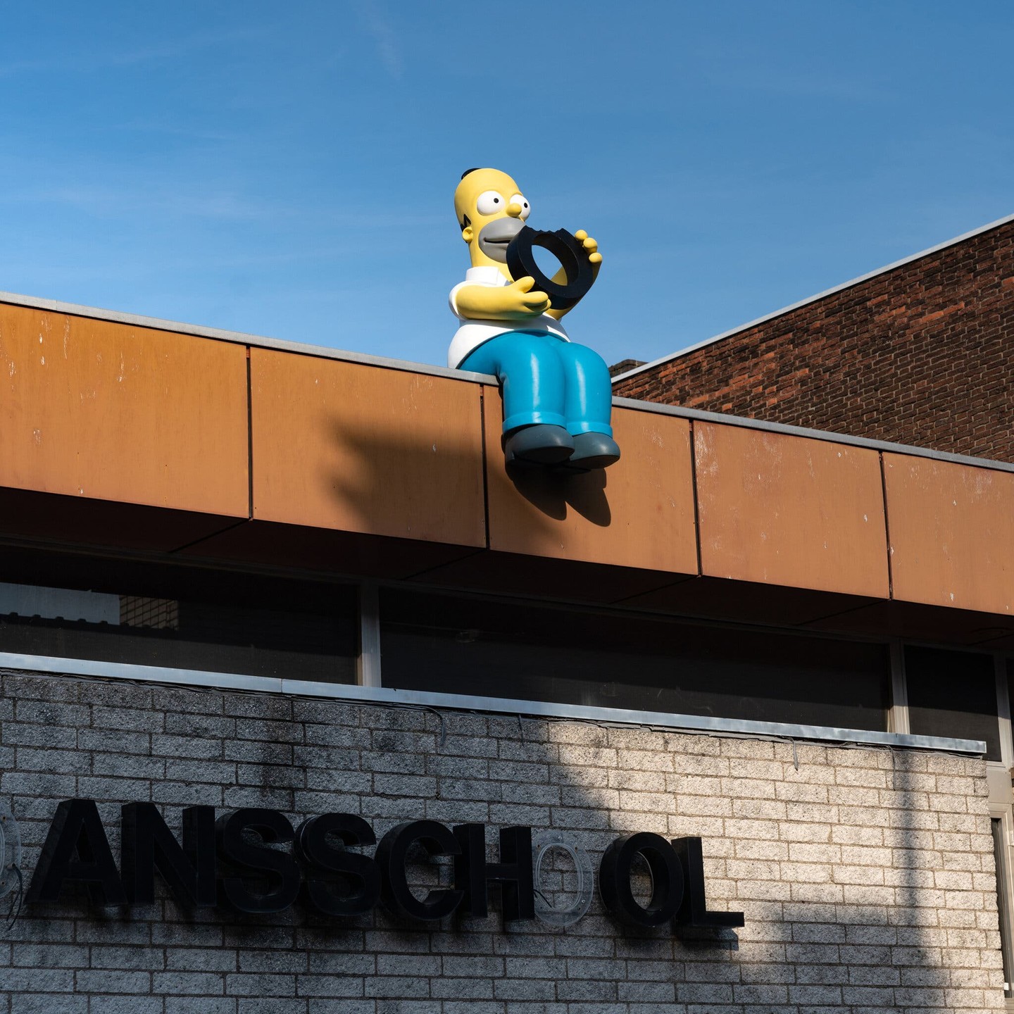 A statue of Homer Simpson sitting on the roof of a school. The letter “O“ has been removed from a sign with the name of the school, and Homer is eating it, like a donut.

Photo by Yvonne Mak