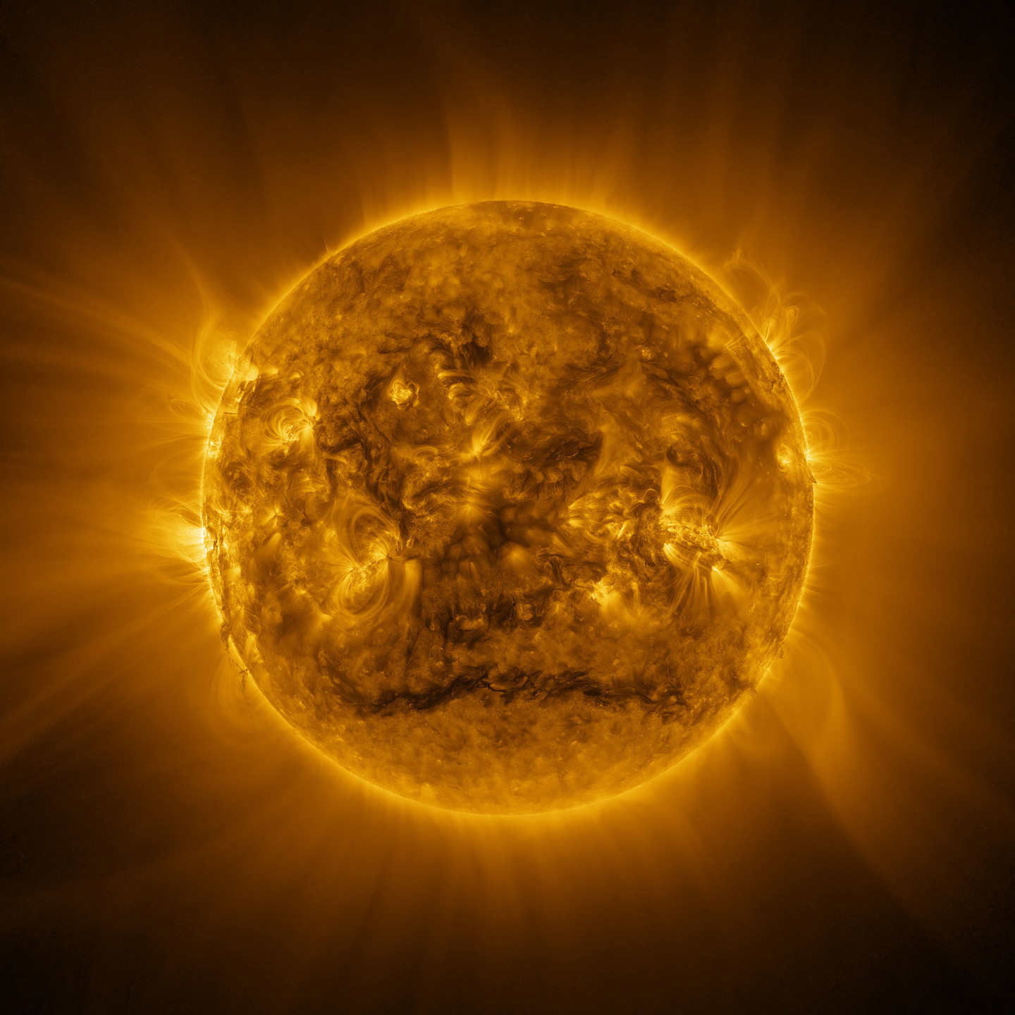 "The Sun looks like a warm yellow sphere with a surface covered with glowing messy hair. The yellow glow extends to the edges of the image, with some regions brighter than others. Many bright yellow arcs stick out from a wide band around the Sun’s equator. A darker region stands out across a roughly horizontal line near the Sun’s south pole. The bright arcs and some darker material can also be seen around the Sun’s edges.